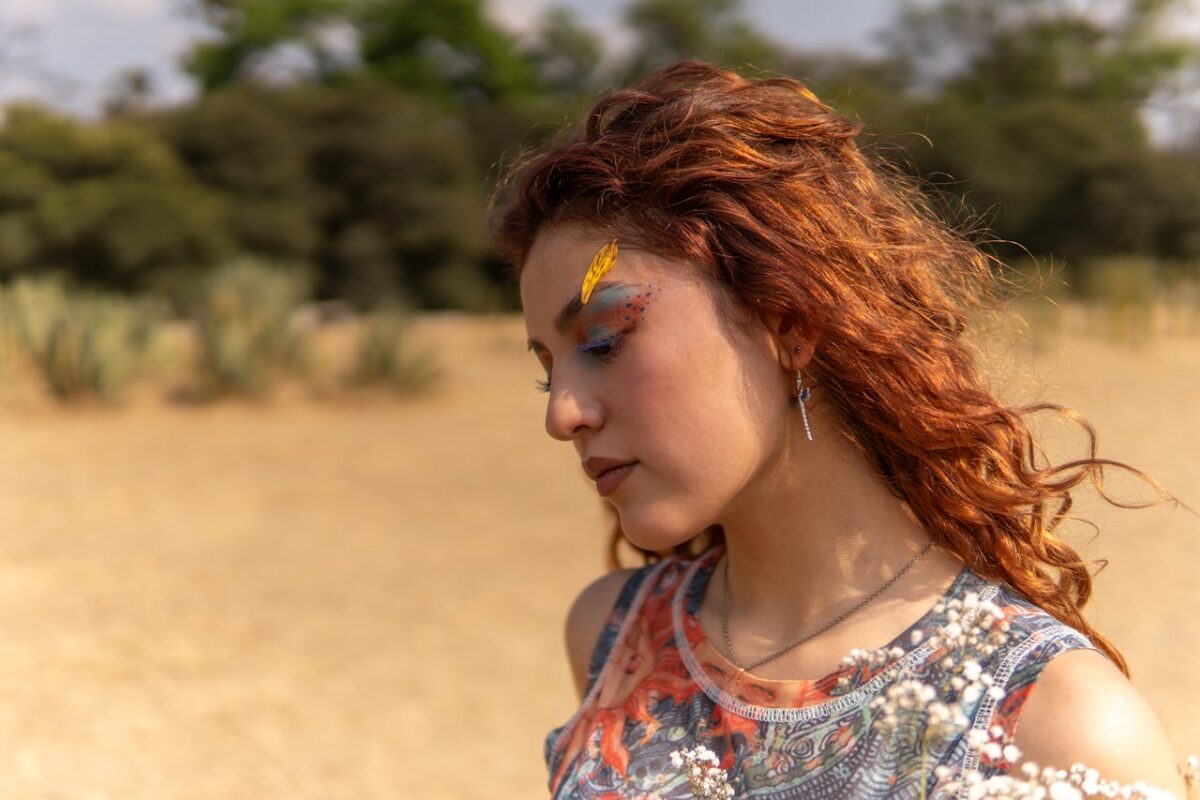 Close-up of a Latina woman with artistic makeup and red hair in a natural setting.