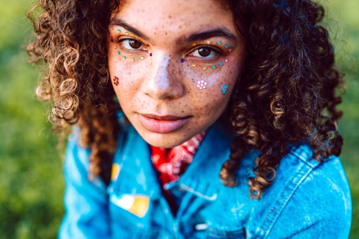 Close-up portrait of a woman with curly hair, freckles, face jewels, and a denim jacket outdoors.